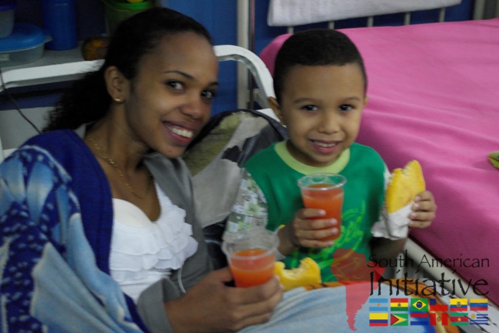 A mother and child smiling with food and juice while sitting together in a hospital setting in Venezuela.