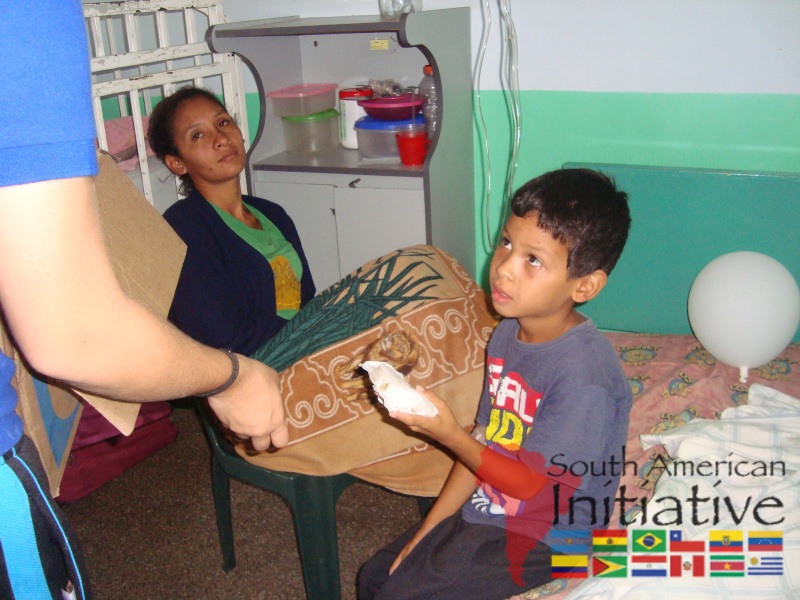 A child receiving food from an SAI volunteer while a caregiver sits nearby in a hospital room in Venezuela.