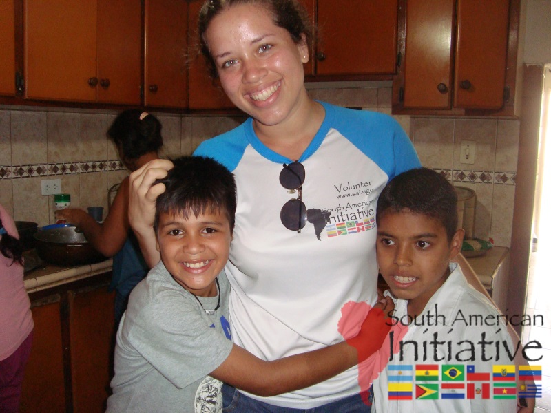 An SAI volunteer smiling with two children inside a family kitchen in Venezuela.