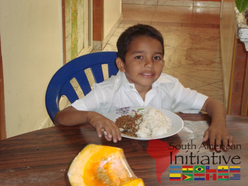 A young boy sitting at a table eating a simple meal with food support connected to SAI in Venezuela.