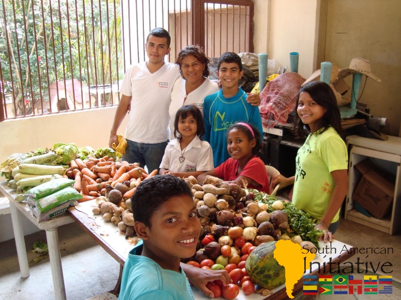 Children and adults gathered around a table of fresh vegetables and produce provided through SAI in Venezuela.