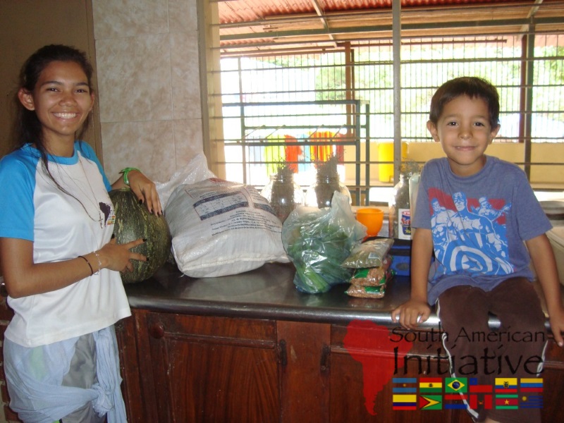 An SAI volunteer standing beside a child with donated groceries and produce in a home in Venezuela.
