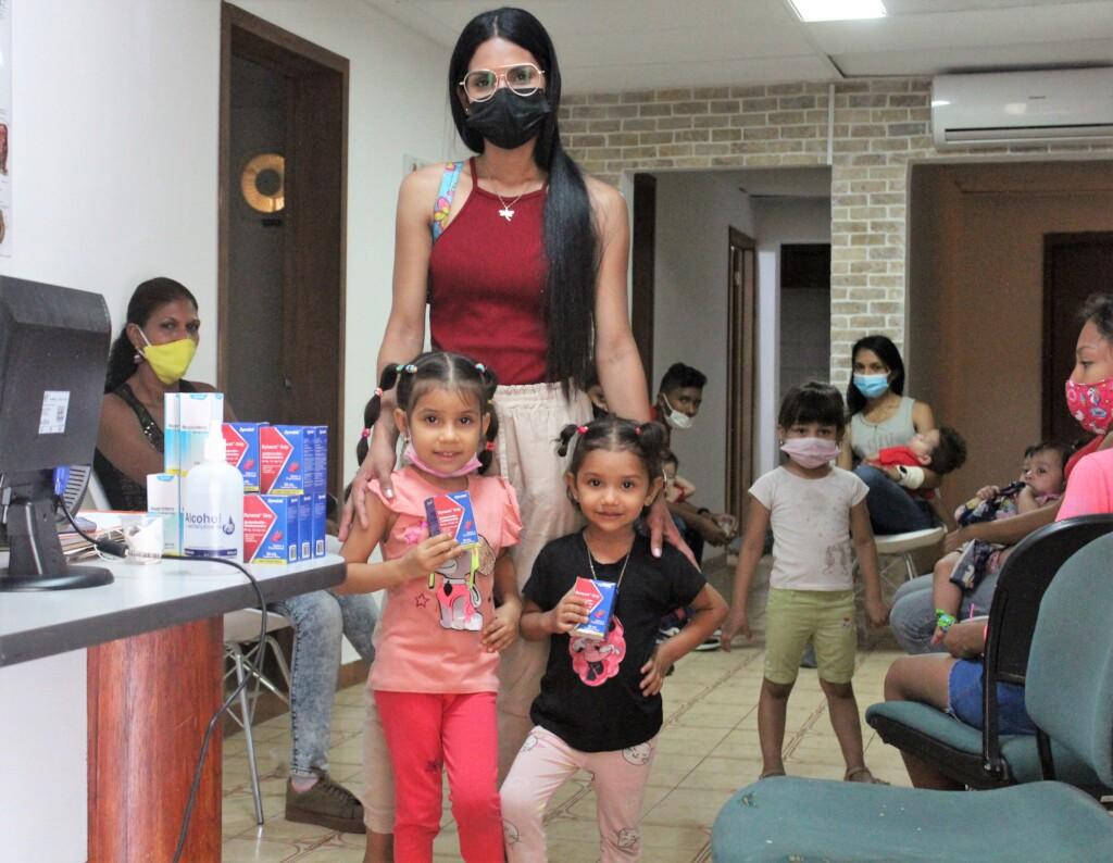 A woman stands with two young girls holding medicine boxes in a clinic waiting room with other children and families nearby.