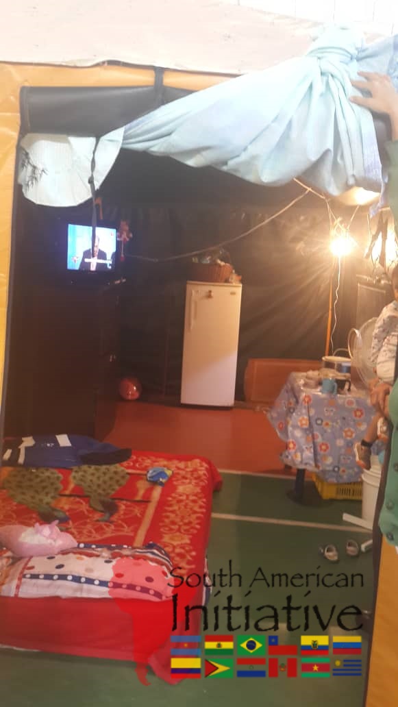 The inside of an emergency shelter tent in Venezuela with bedding, a small refrigerator, and basic living supplies.