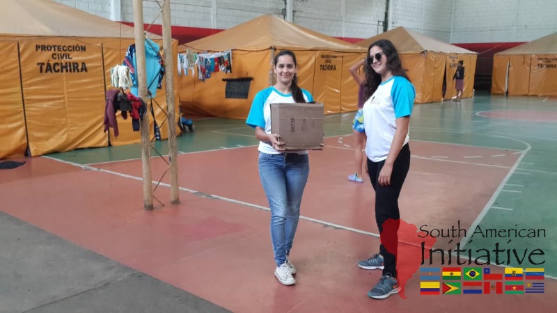 Two SAI volunteers stand inside a refugee shelter in Venezuela, with one carrying a box of supplies.