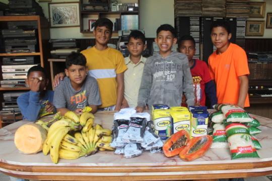 SAI Delivers Nutritious Food Supplies to Support Orphaned Children in Venezuela SAI (South American Initiative), a compassionate non-profit in Venezuela, provides essential nutrition to orphaned children through its food distribution programs. This image captures a meaningful moment where volunteers prepare to distribute fresh produce and packaged food items, including bananas, squash, rice, and tea. The table brimming with healthy options reflects SAI's dedication to combating hunger and ensuring the well-being of vulnerable children. Set against the backdrop of a cozy room filled with books and other resources, the scene embodies the warmth and care brought to each outreach event. Thanks to the tireless efforts of volunteers and the generosity of donors, SAI continues to nourish and uplift the lives of orphaned children across Venezuela.