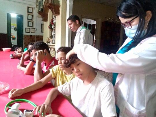 SAI Provides Medical Check-Ups and Nutrition Support to Orphaned Children in Venezuela SAI (South American Initiative), a compassionate non-profit in Venezuela, extends its care to orphaned children through medical check-ups and nutritious food distribution. In this image, a SAI volunteer in a white coat uses a green stethoscope to examine a child seated at a red table. A white cup, plastic containers, and a few other items on the table illustrate the practical setup for these health assessments. The setting includes walls adorned with framed pictures and certificates, creating an inviting indoor environment. This scene exemplifies SAI’s holistic approach to supporting orphaned children by addressing their healthcare and nutritional needs. With the commitment of passionate volunteers and the generosity of donors, SAI continues to bring hope and essential care to vulnerable children across Venezuela.